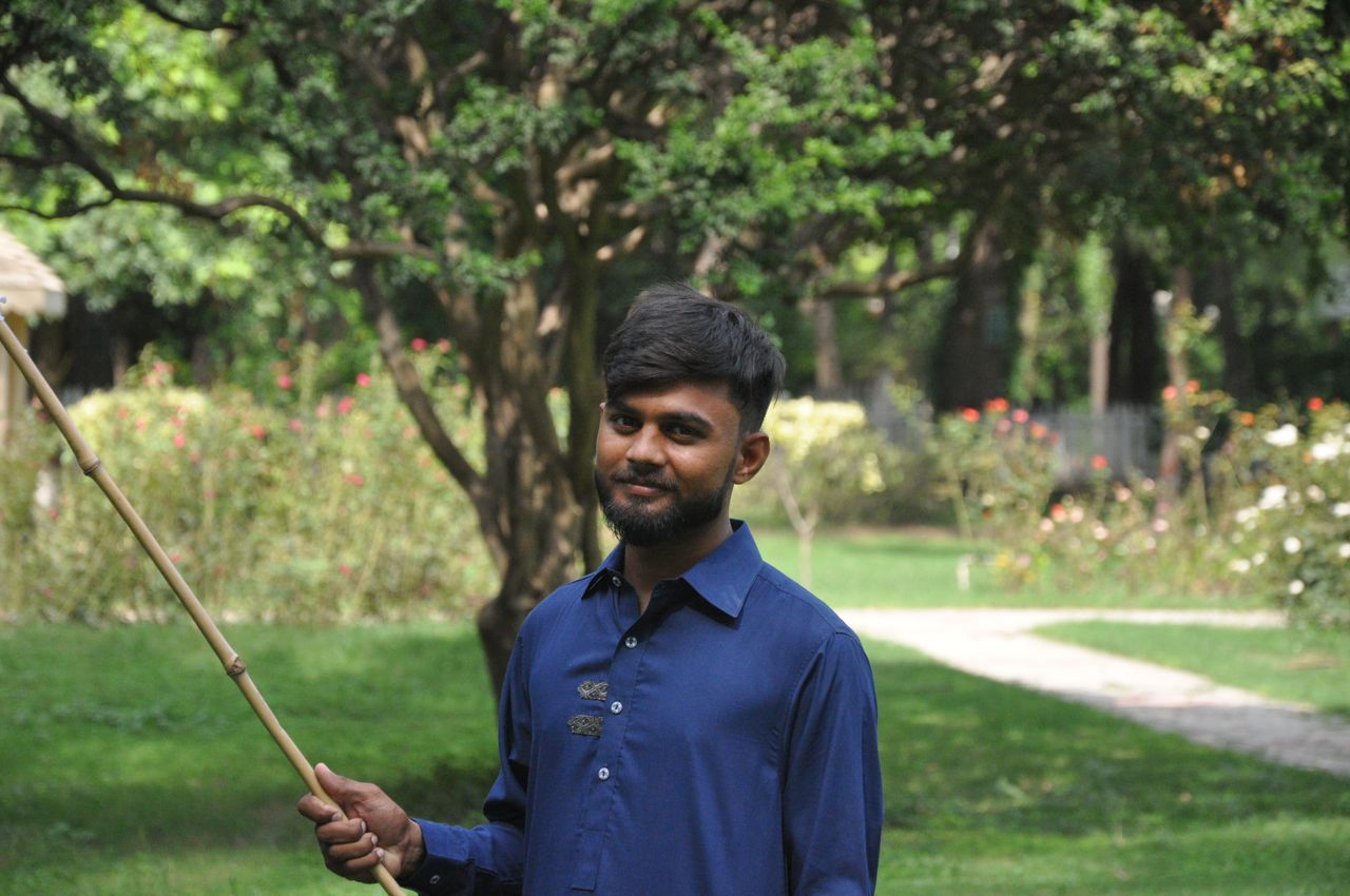 A cheerful man in a blue shirt holding a stick in a sunny green park setting.
