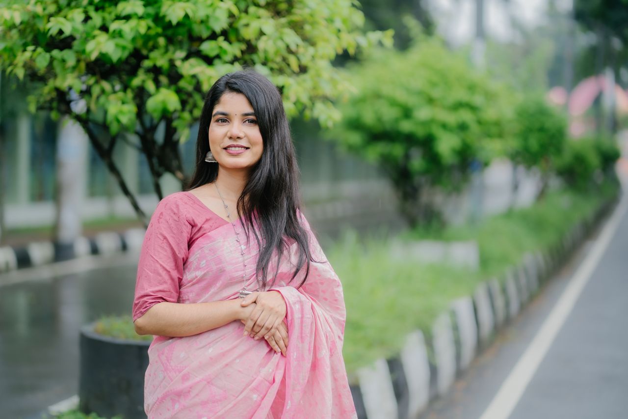 Charming woman wearing a pink saree, standing outdoors with greenery in the background.